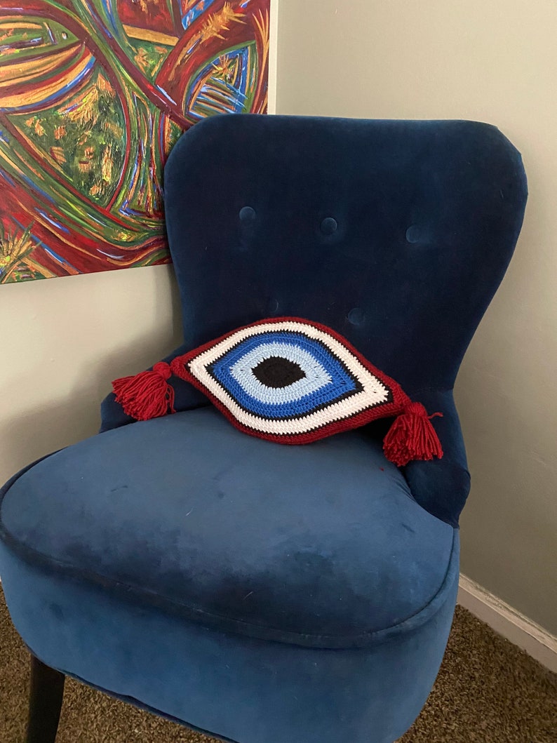 以下が含まれることがあります： 赤い房飾りがついた、青いベルベットの肘掛け椅子に、クロシェで編まれた邪眼のクッションが置かれています。クッションは白、赤、青、黒です。