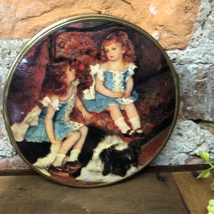May include: A round, brass-framed picture with a vintage image of two girls in blue dresses sitting with a black and white dog. The girls are in a room with a floral patterned armchair.