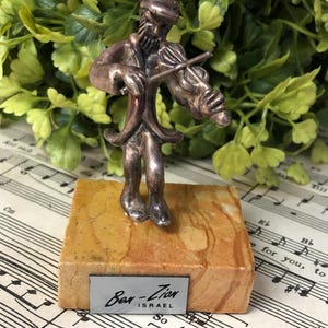 May include: A silver-toned figurine of a violinist playing a violin, mounted on a rectangular, marbled base. The base has a small metal tag that reads "Bar-Zion Israel". The background includes sheet music and greenery.