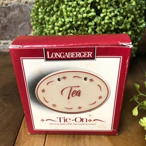 May include: A red and white Longaberger box featuring an oval tea tag. The tag is cream-colored with the word "Tea" in red script and small floral accents. The box reads "Tie-On" and "Trim Your Basket With This Lovely Accessory."