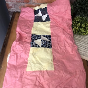 May include: A pink quilt with a central design of yellow and navy blue squares and white star-like shapes. The quilt is folded on a wooden surface, with a brick wall in the background.