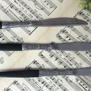 May include: Three butter knives with black handles and silver blades. The silver portion has an etched design. The knives are arranged on a sheet of vintage sheet music.