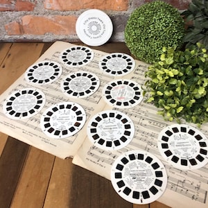May include: A collection of vintage View-Master reels and a white container with text. The reels are circular with small rectangular windows and text. They are arranged on a sheet of vintage sheet music. A green plant and brick wall are in the background.