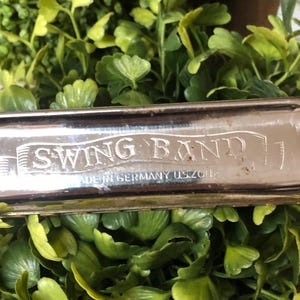 May include: A vintage harmonica with the text "SWING BAND" engraved on the metal surface. The harmonica is silver-toned and appears to be made in Germany. The instrument is set against a backdrop of green foliage.