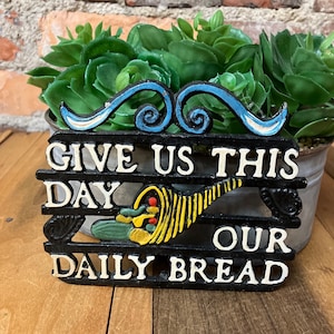 May include: Black cast iron sign with white lettering that reads "Give us this day our daily bread". The sign has a decorative swirl design at the top and a stylized image of a cornucopia.