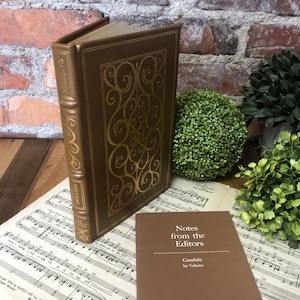 May include: A hardcover book titled "Candide" by Voltaire, with a brown cover and gold decorative accents. The book is displayed with a smaller booklet titled "Notes from the Editors" and sheet music in the background.