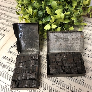 May include: Two rectangular metal boxes containing a collection of small, dark, individual metal letterpress type blocks. Each box is open with the lid upright, revealing rows of stacked, weathered, metallic characters used for printing.