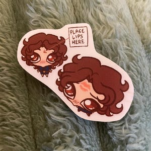 Op de afbeelding: Een gestanste sticker met twee cartoon illustraties van een persoon met bruin krullend haar en grote ogen. Eén illustratie heeft een klein vakje met de tekst "PLACE LIPS HERE". De andere illustratie heeft een kus op de wang.