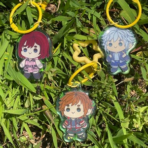 May include: Three acrylic keychains featuring cartoon characters with yellow key rings. The characters have red, blue, and brown hair, wearing colorful outfits. The keychains are set against a background of green grass.