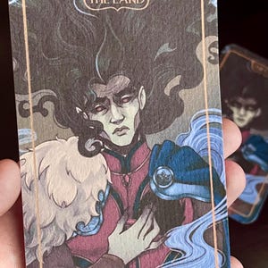 Puede incluir: Ilustración de carta de tarot con una figura de pelo largo y oscuro y expresión seria. La carta está enmarcada en oro con las palabras "THE LAND" en la parte superior. La figura viste un atuendo rojo y azul, con una criatura peluda blanca a la izquierda.