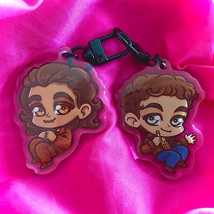 Puede incluir: Dos llaveros con personajes de dibujos animados, uno con cabello castaño y una chaqueta marrón, el otro con cabello castaño y una chaqueta azul. Ambos personajes tienen ojos marrones y sonríen.