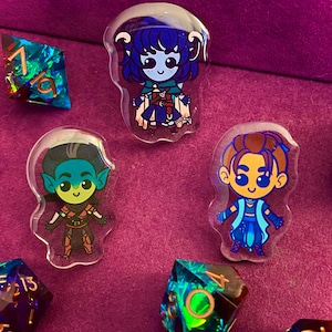May include: Three character pins featuring cartoon illustrations of fantasy characters. The pins are clear with a white background and feature a blue, green, and brown character with a blue, green, and brown background.
