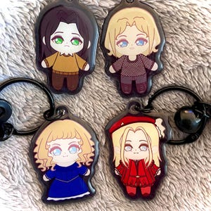 May include: Four anime-style keyrings with black fixings. Each keyring features a different character with large eyes and unique outfits. The characters have blonde, brown, and red hair. The outfits include a red coat, blue dress, and brown sweater.
