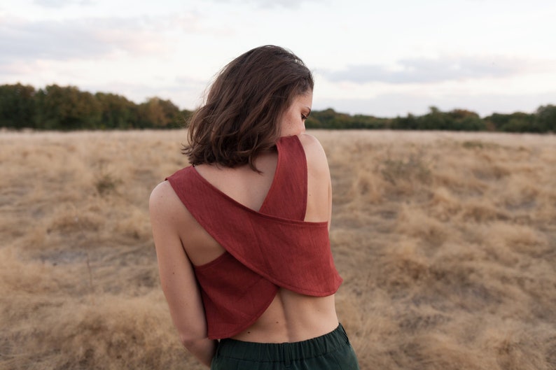 Op de afbeelding: Een vrouw draagt een roestkleurig linnen topje met een gekruist rugontwerp. Het topje is kort en heeft een ontspannen pasvorm. De vrouw staat in een veld met droog gras.