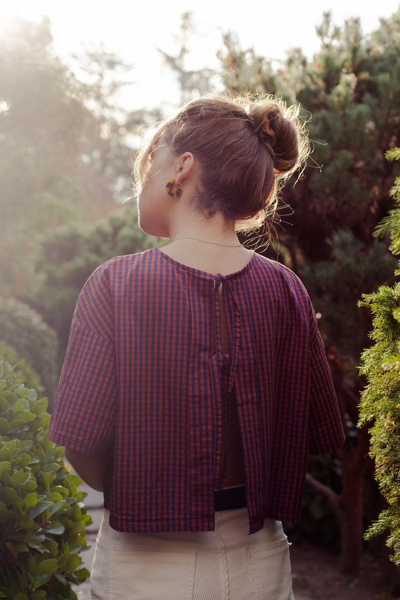 Op de afbeelding: Een vrouw draagt een rood-blauw geruit overhemd met een strik op de rug. Het overhemd is getailleerd en heeft korte mouwen. Ze draagt een witte broek en heeft haar haar in een knotje.