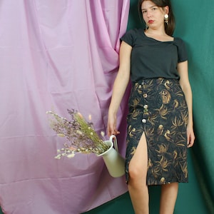 Peut inclure: Une femme portant un t-shirt noir et une jupe marron foncé à imprimé doré et fente sur le côté. Elle se tient devant un rideau rose et un mur vert. Elle tient un pichet blanc avec des fleurs séchées.