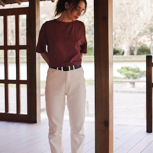 Op de afbeelding: Een vrouw in een rood-wit geruit overhemd, witte jeans en zwarte schoenen staat in een houten constructie.