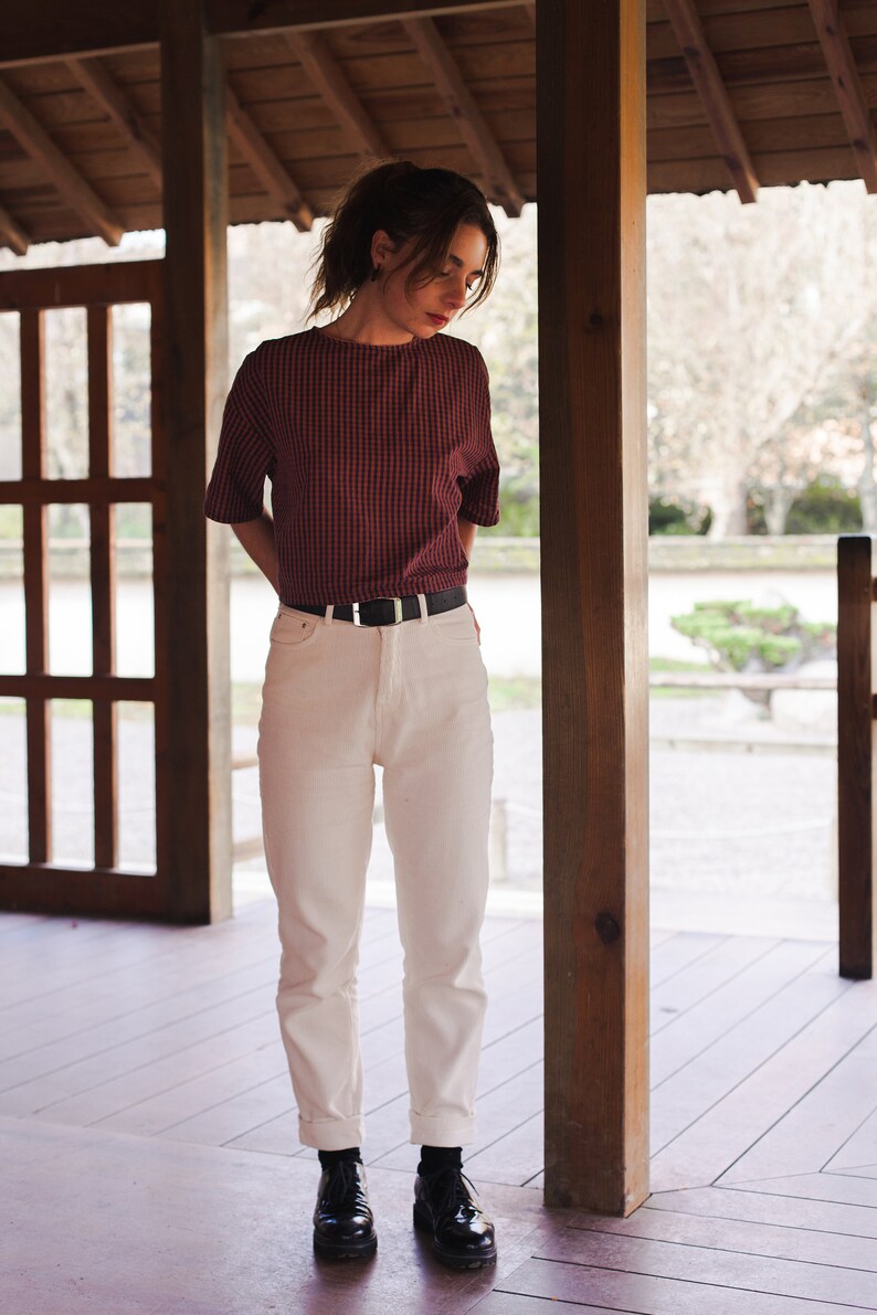 Op de afbeelding: Een vrouw in een rood-wit geruit overhemd, witte jeans en zwarte schoenen staat in een houten constructie.