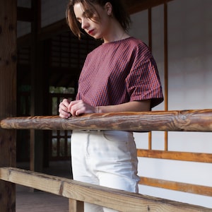 Op de afbeelding: Een vrouw in een rood-blauw geruit overhemd en een witte broek staat op een houten dek. Ze leunt tegen een houten railing.