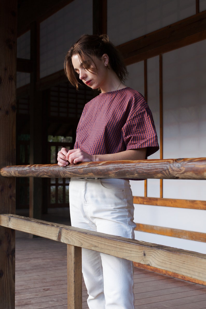 Op de afbeelding: Een vrouw in een rood-blauw geruit overhemd en een witte broek staat op een houten dek. Ze leunt tegen een houten railing.