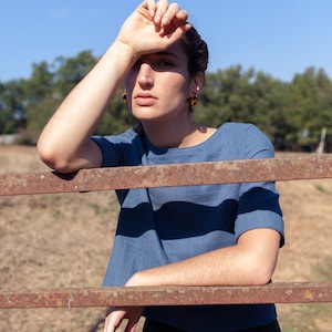 Op de afbeelding: Een vrouw die een blauw linnen top draagt met een ronde hals en korte mouwen, staand voor een roestig metalen hek. Ze draagt een groene broek en heeft haar hand op haar voorhoofd.