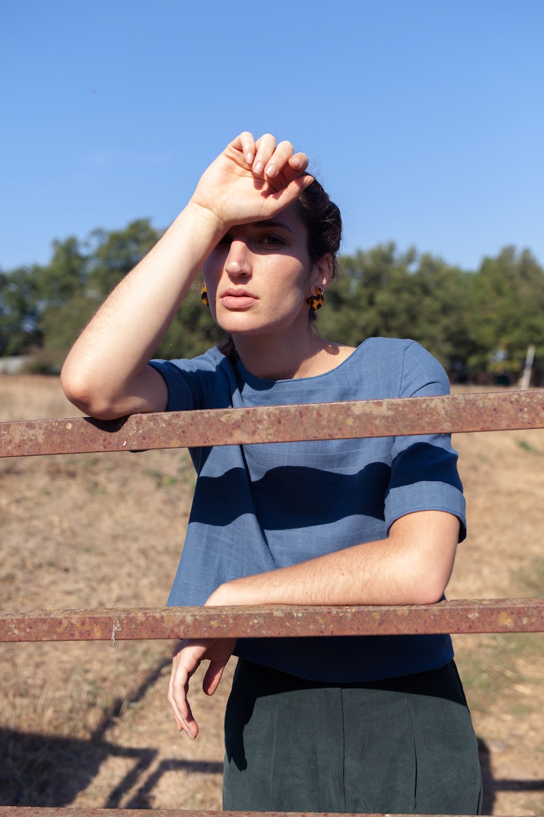 Op de afbeelding: Een vrouw die een blauw linnen top draagt met een ronde hals en korte mouwen, staand voor een roestig metalen hek. Ze draagt een groene broek en heeft haar hand op haar voorhoofd.
