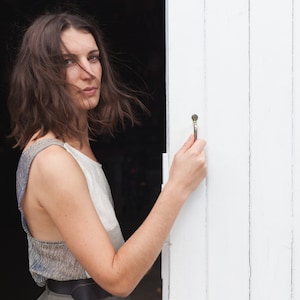 Op de afbeelding: Een vrouw met lang bruin haar staat voor een witte houten deur. Ze draagt een lichtgrijs, mouwloos topje en een donkerbruine riem. Ze houdt de deurknop vast met haar rechterhand.