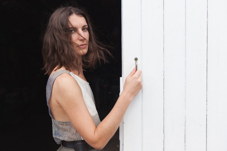 Op de afbeelding: Een vrouw met lang bruin haar staat voor een witte houten deur. Ze draagt een lichtgrijs, mouwloos topje en een donkerbruine riem. Ze houdt de deurknop vast met haar rechterhand.