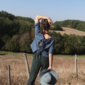 Op de afbeelding: Een vrouw in een blauw shirt met een veter aan de achterkant en een donkergroene broek staat in een veld en houdt een blauw-witte strohoed vast.