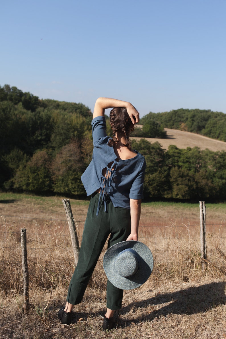 Op de afbeelding: Een vrouw in een blauw shirt met een veter aan de achterkant en een donkergroene broek staat in een veld en houdt een blauw-witte strohoed vast.