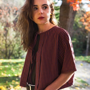 Op de afbeelding: Een vrouw draagt een rood-blauw geruit overhemd met een witte kraag en een witte riem. Ze staat in een park met herfstbladeren op de achtergrond.