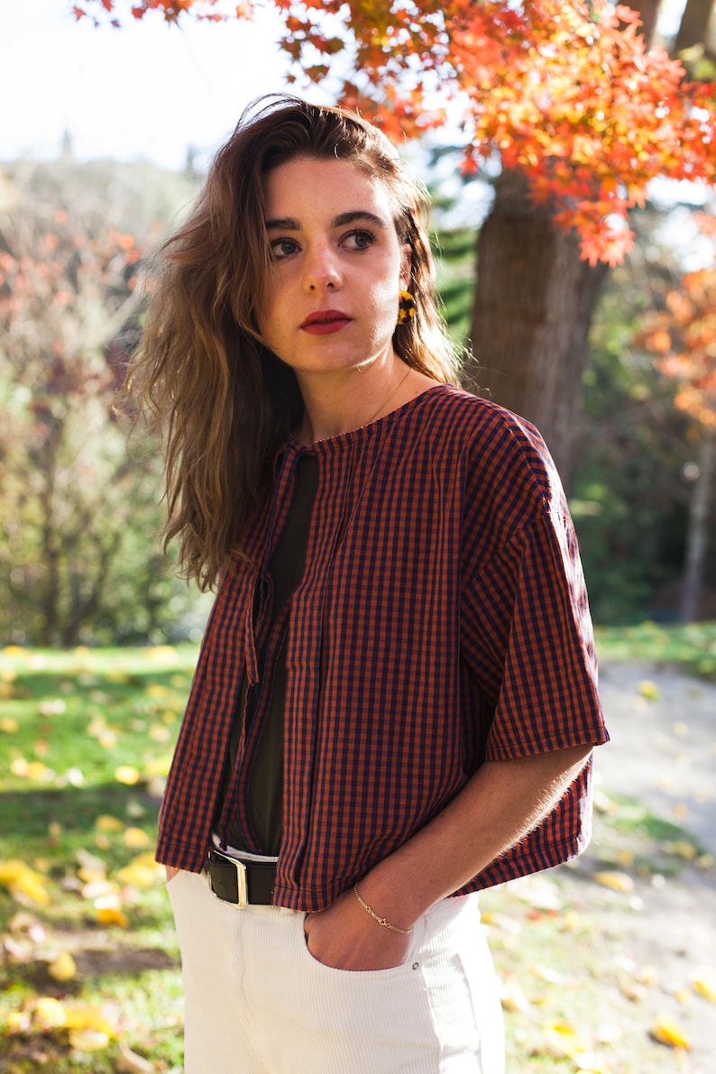 Op de afbeelding: Een vrouw draagt een rood-blauw geruit overhemd met een witte kraag en een witte riem. Ze staat in een park met herfstbladeren op de achtergrond.