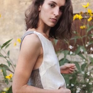 Op de afbeelding: Een vrouw met bruin haar draagt een wit, mouwloos topje met een grijs en bruin patroon. Ze staat voor een muur met gele bloemen.