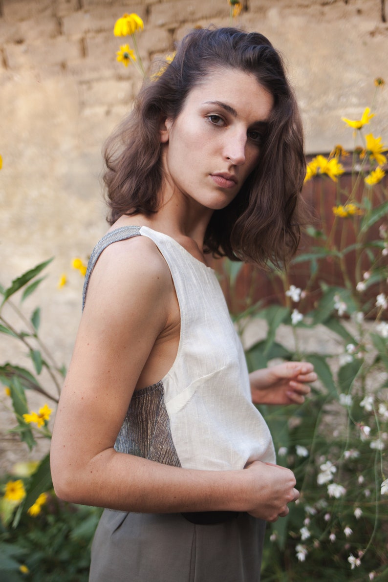 Op de afbeelding: Een vrouw met bruin haar draagt een wit, mouwloos topje met een grijs en bruin patroon. Ze staat voor een muur met gele bloemen.