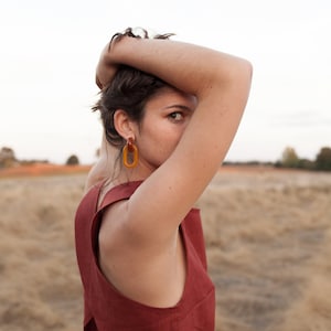 Op de afbeelding: Een vrouw draagt een roestkleurig, mouwloos topje met een uniek asymmetrisch ontwerp. Ze draagt grote, ovale amberen oorbellen.