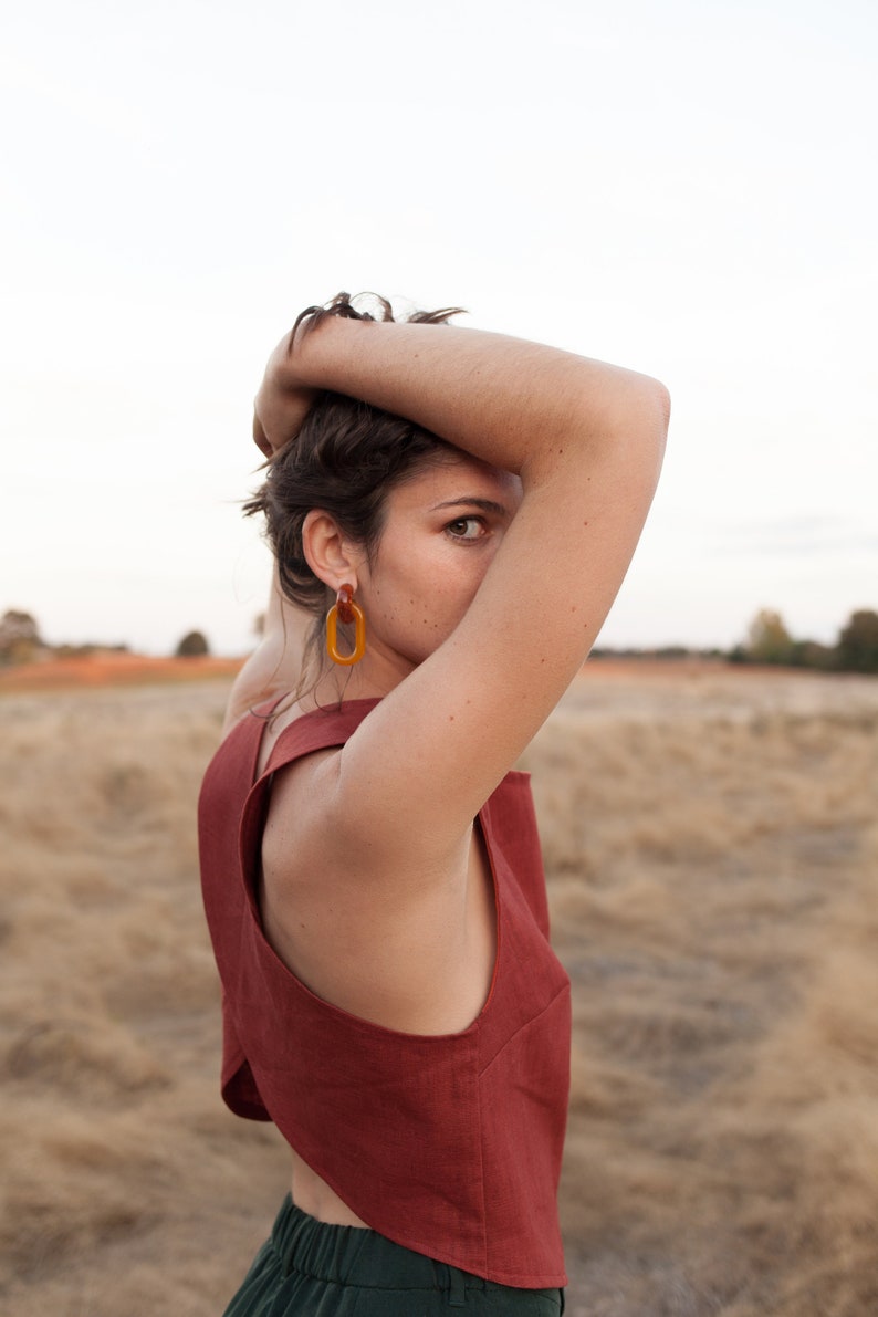 Op de afbeelding: Een vrouw draagt een roestkleurig, mouwloos topje met een uniek asymmetrisch ontwerp. Ze draagt grote, ovale amberen oorbellen.