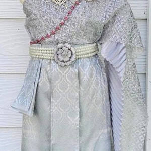 May include: A silver Thai traditional dress with a white flower on the belt, a silver necklace, and a white flower crown.
