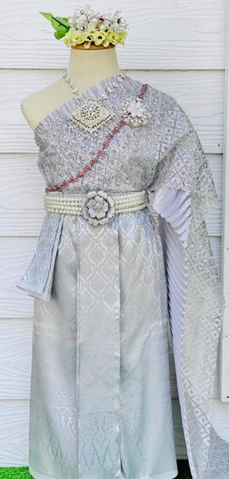 May include: A silver Thai traditional dress with a white flower on the belt, a silver necklace, and a white flower crown.