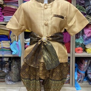 May include: A gold-colored traditional Thai outfit for a child. The outfit includes a short-sleeved shirt, pants, and a decorative sash. The sash is dark brown with gold accents and a jeweled clasp. The pants and sash have an ornate pattern.