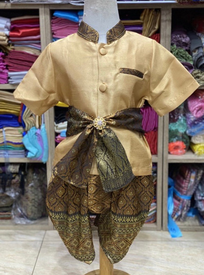 May include: A gold-colored traditional Thai outfit for a child. The outfit includes a short-sleeved shirt, pants, and a decorative sash. The sash is dark brown with gold accents and a jeweled clasp. The pants and sash have an ornate pattern.