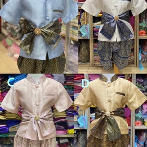May include: Four traditional Thai children's outfits in various colors: light blue, white, pink, and tan. Each set includes a short-sleeved shirt, patterned pants, and a decorative sash with a central embellishment. The outfits are displayed on mannequins.