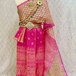 May include: A pink and gold traditional Thai dress with a gold patterned fabric and a gold belt with green jewels. The dress is on a mannequin and has a sheer pink fabric draped over the shoulders.