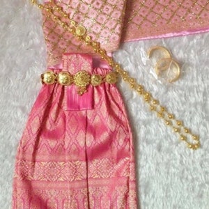 May include: A pink and gold traditional Thai dress with a matching sash, necklace, and bracelets. The dress has a patterned design and is decorated with gold accents.