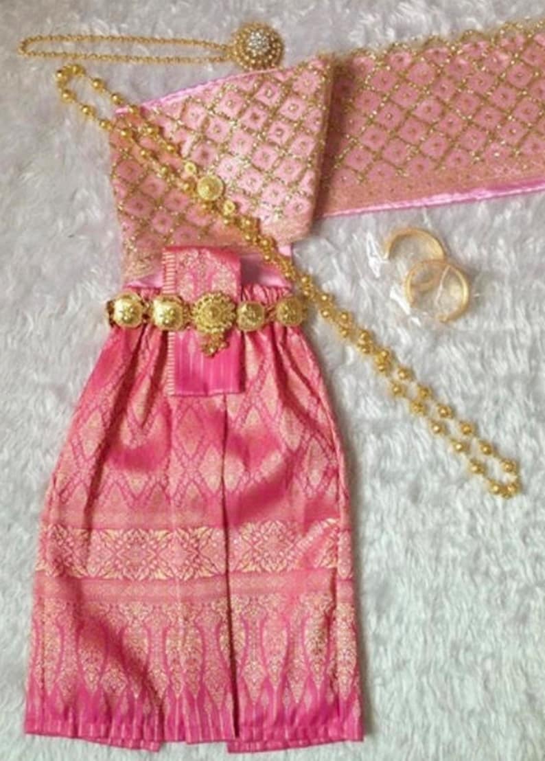 May include: A pink and gold traditional Thai dress with a matching sash, necklace, and bracelets. The dress has a patterned design and is decorated with gold accents.
