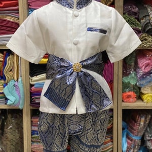 May include: A traditional Thai outfit for a child, featuring a white short-sleeved shirt with a mandarin collar and decorative blue trim. The outfit includes matching blue patterned shorts and a decorative sash with a gold-colored embellishment.