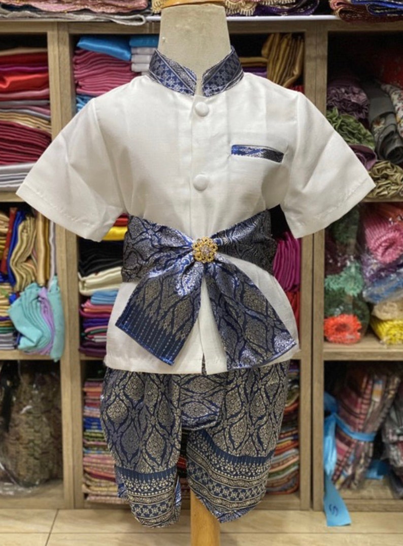 May include: A traditional Thai outfit for a child, featuring a white short-sleeved shirt with a mandarin collar and decorative blue trim. The outfit includes matching blue patterned shorts and a decorative sash with a gold-colored embellishment.