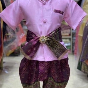 May include: A traditional Thai outfit for children. The ensemble features a light pink short-sleeved shirt with a mandarin collar and matching pocket detail. The outfit includes patterned purple pants and a decorative sash with a gold-colored buckle.