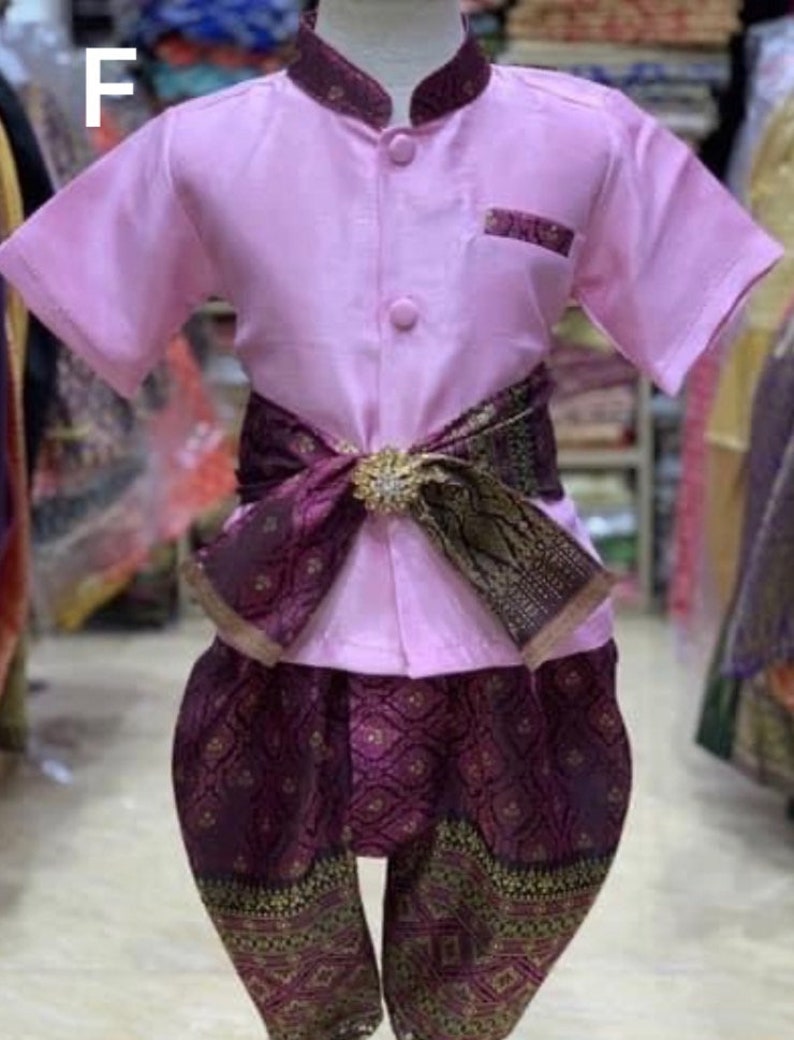 May include: A traditional Thai outfit for children. The ensemble features a light pink short-sleeved shirt with a mandarin collar and matching pocket detail. The outfit includes patterned purple pants and a decorative sash with a gold-colored buckle.