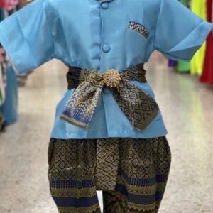May include: A light blue traditional Thai outfit for children. The outfit includes a short-sleeved shirt, patterned pants, and a decorative belt. The shirt has a small pocket and a mandarin collar. The pants and belt feature a gold and navy blue pattern.