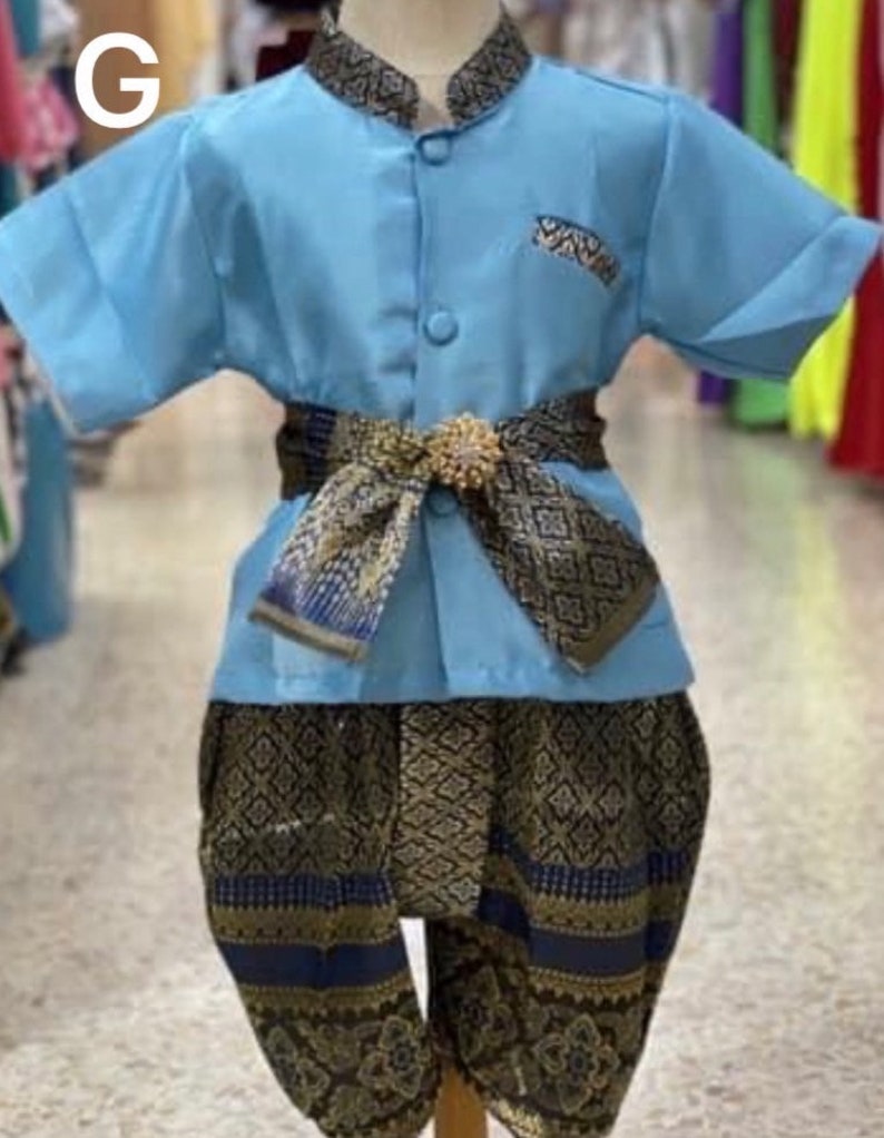 May include: A light blue traditional Thai outfit for children. The outfit includes a short-sleeved shirt, patterned pants, and a decorative belt. The shirt has a small pocket and a mandarin collar. The pants and belt feature a gold and navy blue pattern.